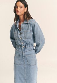 Pitkähihainen farkkupaitamekko, jossa on istuva vyötärö, kaksi rintataskua, napit ja vaaleansininen pesu.