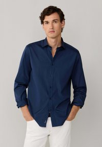 Granatowa koszula z guzikami o gładkiej fakturze, z długimi rękawami i czarnymi guzikami; włożona w jasne spodnie; casualowy fason.