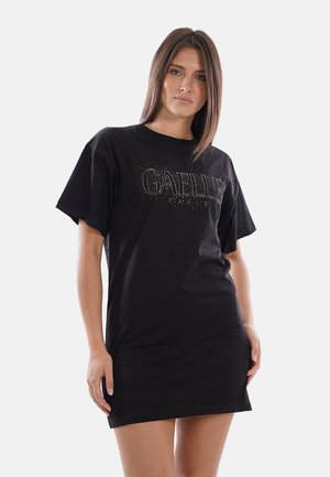 Donna con capelli castani lisci indossa un vestito T-shirt nero oversize con il logo "GAËLLE PARIS" in strass sul davanti.