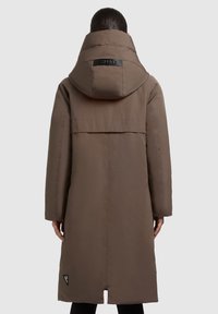 Bruine lange parka met een grote capuchon, voorzien van een logopatch op de achterkant, een rits aan de voorkant en een recht model met zijzakken.