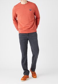 Sweatshirt in roestkleur met een ronde hals en een klein blauw logo op de borst, gecombineerd met grijze jeans en bruine veterschoenen.