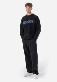 Sweater preto com texto em azul com franjas na frente, combinado com calças pretas de corte largo e ténis brancos. Conjunto simples e casual.
