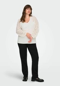 Hvit bluse med lange ermer i blonder, v-hals og løs passform, kombinert med svarte bukser. Skoene er svarte med en glatt finish.