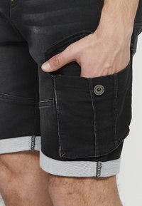Shorts de mezclilla negra con dobladillo enrollado, bolsillo utilitario, acentos de costura amarilla y detalle de botón metálico. Presenta un diseño de ajuste slim.