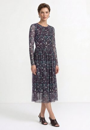 Donna che indossa un vestito midi a maniche lunghe con motivo floreale scuro e scarpe nere aperte con tacco, in piedi su uno sfondo bianco uniforme.