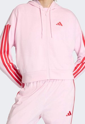 Personne portant un survêtement rose clair avec des rayures rouges sur les manches et les pantalons, ainsi qu'un petit logo rouge sur la poitrine et la cuisse.