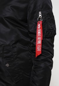 Czarna bomberka z gładkiego nylonu, z zamkiem błyskawicznym na rękawie oraz czerwoną etykietą z napisem "USUNĄĆ PRZED STARTEM".