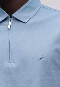 Κοντινή λήψη ανοιχτό μπλε πόλο με φερμουάρ και μικρό κεντητό λογότυπο "MK" στο στήθος, φορεμένο από άτομο με σκουρόχρωμο τόνο δέρματος.