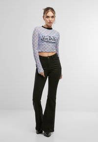 Langermet cropped topp i lys blått, gjennomsiktig stoff med gjentatt logo-mønster, kombinert med svarte vide jeans og svarte støvler.