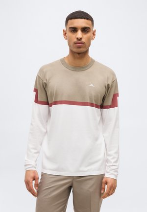 Mand iført en beige, hvid og rød langærmet skjorte med et lille logo på brystet, stående mod en ensartet lys baggrund.