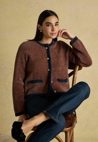 Brūns teksturēts kardigans ar tumši zilām apmalēm, apaļu kakla izgriezumu un zeltītām pogām; ir divas tumši zilas kabatas. Modelis sēž uz koka krēsla.