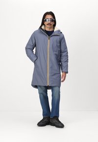 Cappotto leggero, grigio, di lunghezza media con dettagli in giallo alla zip, foderato in morbido tessuto e dotato di cappuccio. Indossato sopra jeans scuri e sneaker nere.