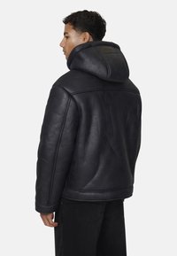 Giacca a vento nera in pelle con cappuccio, caratterizzata da una consistenza morbida, una vestibilità rilassata e dettagli cuciti, con colletto alto e design minimalista.