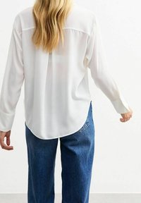 Blusa bianca a maniche lunghe con una texture liscia, orlo arrotondato e polsini con bottoni, abbinata a jeans blu larghi.