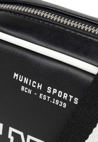 Svart sportväska med vita accenter, med en silverdragkedja och tryckt text: "MUNICH SPORTS BCN - EST. 1939." Texten har ett modernt typsnitt.