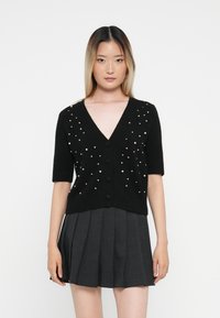 Svart kortermet cardigan med V-hals, dekorert med spredte hvite perler og fem svarte knapper langs fronten.