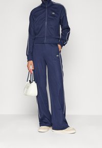 Chándal azul marino de tejido suave, con rayas laterales blancas, una chaqueta con cremallera y cuello alto, y pantalones de corte holgado. Bolso blanco.