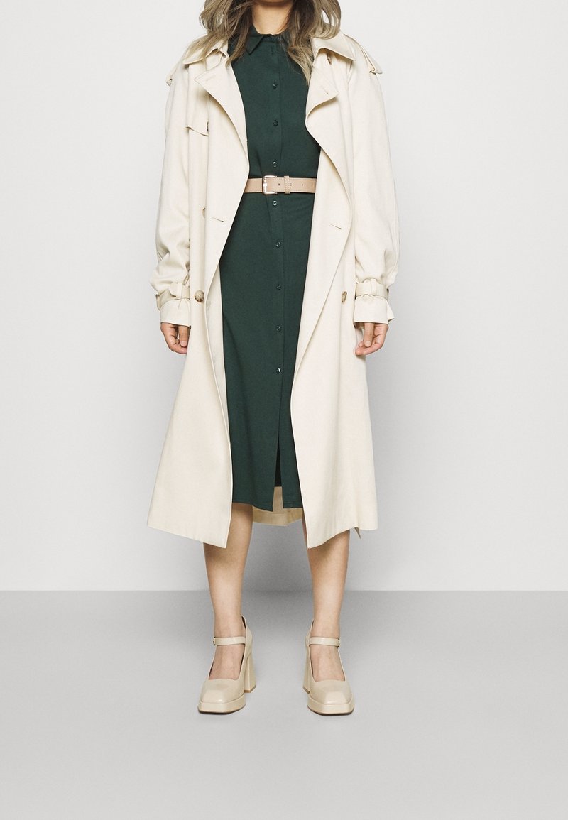 Femme portant une robe boutonnée vert foncé avec une ceinture beige, un long trench-coat beige et des chaussures Mary Jane beige à talons hauts.