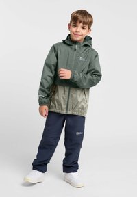 Grüne und beige Kapuzenjacke mit elastischen Bündchen, Reißverschluss und Logo; kombiniert mit marineblauen Hosen und weißen Sneakers. Glattes, leichtes Material.