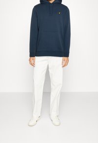 Felpa navy con tasca frontale e cappuccio, abbinata a pantaloni bianchi e sneakers di colore chiaro. Presenta un logo ricamato sul petto.