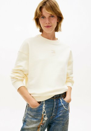 Jeune personne aux cheveux courts portant un sweat crème et un jean bleu avec des accessoires en chaîne, les mains dans les poches, debout devant un fond blanc.