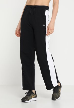 Zwarte wijde broek met witte zijpanelen en klein logo, gedragen met zwarte en gouden sportschoenen, getoond op een staand persoon.