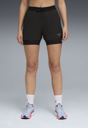 Person iført sorte lagdelte shorts med "HYROX"-logo, hvide mellemlange sokker og grå atletikskotøj med lyserøde og sorte detaljer.