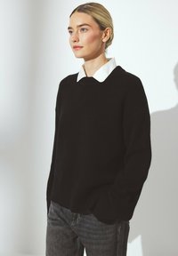 Black sweater with a rounded neckline, ribbed cuffs and hem, layered over a white collared shirt. Background is softly lit.