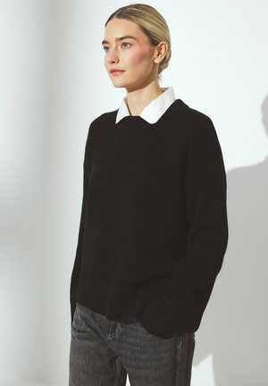 Black sweater with a rounded neckline, ribbed cuffs and hem, layered over a white collared shirt. Background is softly lit.