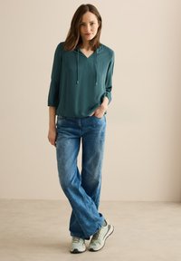 Teal langærmet top med V-hals og snor, parret med lyseblå vide jeans og lysegrønne sneakers. Glat stofstruktur.