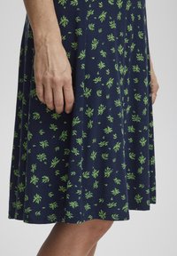 Námořnické šaty s potiskem zelených listů, vyrobené z měkké tkaniny. Mají rozšířený střih, délku ke kolenům a jemné švy.