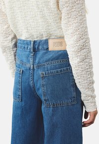 Blauwe denim jeans met een hoge taille, met twee achterzakken en een beige leren label met de tekst "KOTON KIDS." Textuur van een witte trui zichtbaar.