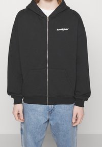 Sudadera con cremallera negra hecha de una mezcla de algodón, con un bolsillo frontal, puños de canalé y un pequeño logo blanco en el pecho.