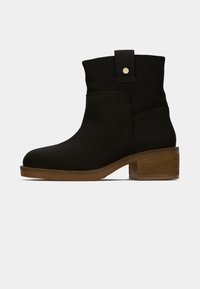 Bottine en suede noire avec un bout arrondi, un talon carré et une semelle de couleur marron clair contrastante. Comprend un détail de bouton pression sur le col.