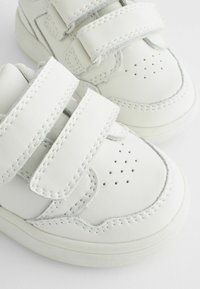 Witte leren sneakers met twee klittenbandsluitingen, geperforeerd te ontwerp en contrasterende stiksels. Zachte textuur en afgeronde vorm.