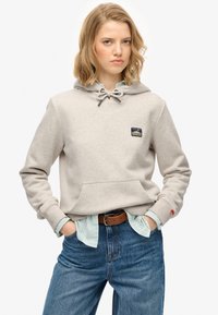 Felpa con cappuccio grigio chiaro con tasca frontale, coulisse regolabile e patch con logo. Indossata sopra una camicia a righe blu e bianche, abbinata a jeans blu.