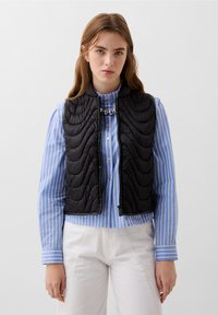 Gilet senza maniche trapuntato nero con collo alto, dotato di chiusura con zip e dettagli di cucitura curva, indossato sopra una camicia a righe blu.