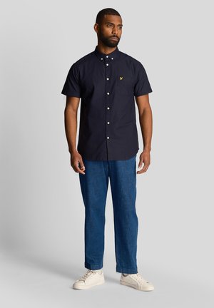 Camicia a maniche corte blu navy con taschino frontale, bottoni bianchi e un piccolo logo giallo; abbinata a jeans blu e sneakers bianche.