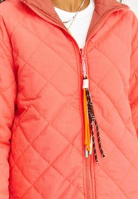 Kurtka w kolorze koralowym z pikowanym wzorem i kontrastowym brązowym kołnierzem; ma zamek błyskawiczny z przodu oraz kolorowe linki do łatwego chwytania.
