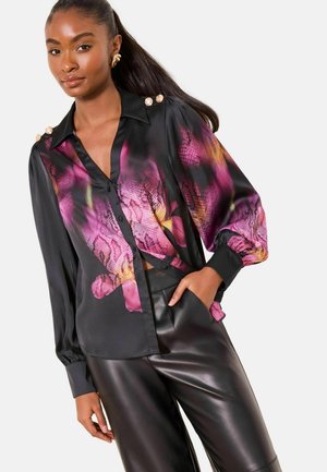 Schwarze Satinbluse mit einem bunten Blumen- und Schlangenprint, ausgestattet mit einem Kragen und goldenen Schulterknöpfen. Kombiniert mit schwarzen Lederhosen.