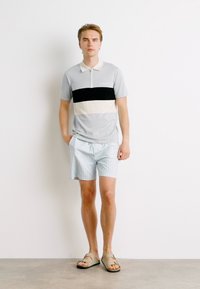 Camisola polo cinza com riscas pretas e brancas, combinada com calções risca de giz azul claro. Usada com sandálias castanhas, apresentada contra um fundo simples.