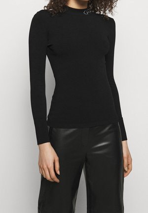 Vrouw draagt een nauwsluitend zwart top met lange mouwen en leren-look zwarte broek tegen een effen lichte achtergrond.