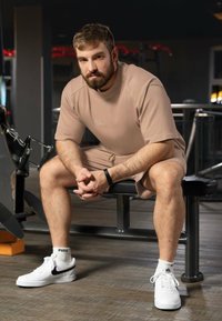 Completo sportivo beige composto da una maglietta a maniche corte e pantaloni corti. Indossato con sneakers bianche Nike e calze a fantasia. Il soggetto è seduto.