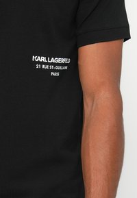 Crna pamučna majica s bijelim natpisom: "KARL LAGERFELD 21 RUE ST-GUILLAUME PARIS." Kratki rukavi i okrugli izrez.