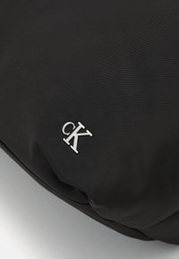 Musta nailonlaukku, jossa on sileä pinta ja hopeanvärinen "CK"-logo. Muotoilu on minimalistinen, pyöristetyin rejoin ja taitettava.