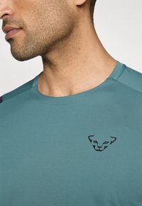 Teal Sportshirt mit strukturierter Oberfläche, ausgestattet mit einem schwarzen Logo auf der Brust und einem runden Halsausschnitt mit kontrastierenden Schulterakzenten.