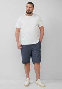 Biała koszulka z krótkim rękawem, granatowe spodenki w paski i kremowe sneakersy noszone przez mężczyznę stojącego na jednolitym tle. Prosty design, swobodny fason.