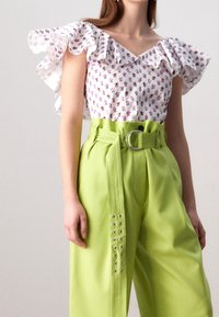 Blusa floreale bianca con maniche arricciate, abbinata a pantaloni verde brillante a vita alta con fibbia e bottoni argentati decorativi.