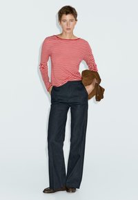 Vrouw in een rood-wit gestreepte langmouwtop, donkere high-waisted broek, met een bruine jas in haar hand, staand tegen een effen lichte achtergrond.