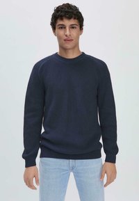 Marineblauer gerippter Strickpullover mit Rundhalsausschnitt, langen Ärmeln und strukturiertem Stoff. Kombiniert mit hellblauen Jeans, präsentiert vor einem einfarbigen Hintergrund.